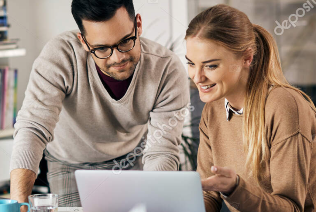 Two people in casual attire are looking at a laptop screen together, deeply engaged in discussing marketing strategies. The setting is a cozy indoor space with books and a plant visible in the background.