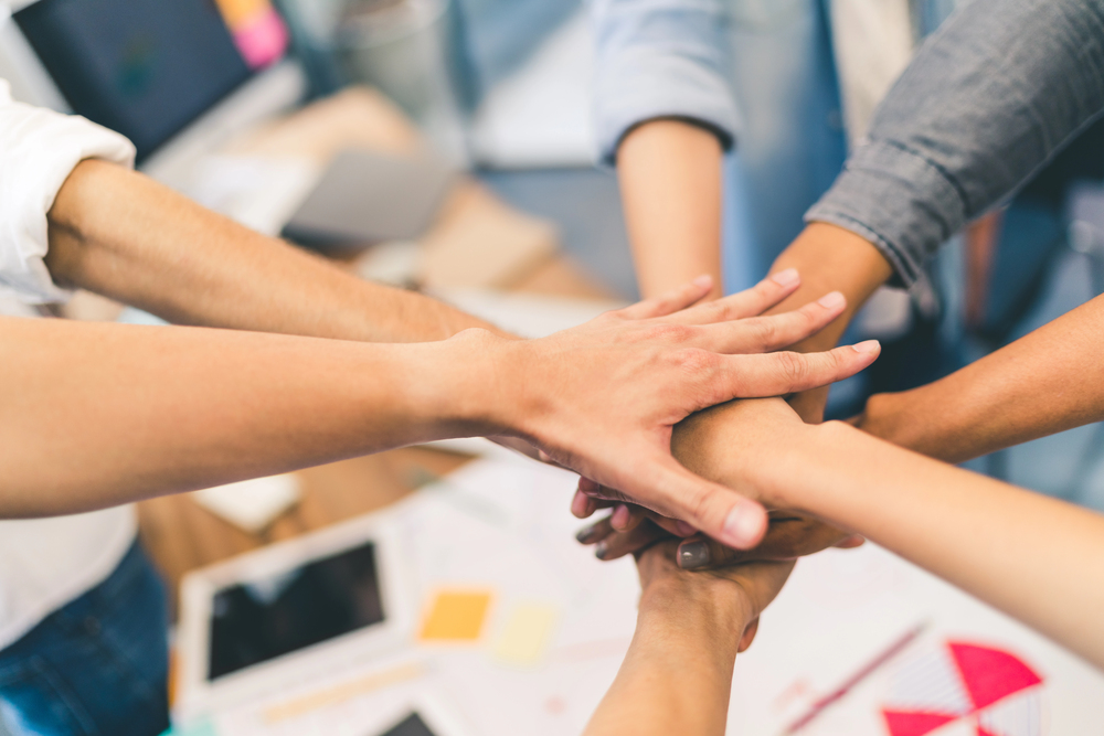 A diverse group of people join hands in a collaborative gesture over a desktop with papers and a tablet. The image conveys teamwork and unity in a professional setting.