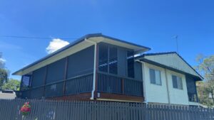 A two-story house with a modern, dark gray exterior on an elevated wooden deck. It features large windows with shutters and outdoor blinds by QLD Shade