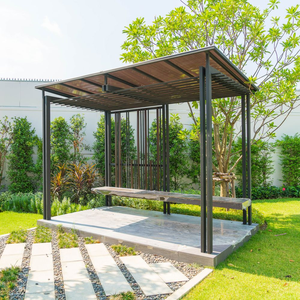 A modern gazebo with a flat roof and metal frame is situated in a landscaped garden. It features a wooden bench and is surrounded by lush greenery, including a tall tree and a neatly trimmed hedge. Stone pathways, grass, and the perfect patio elements complete this serene outdoor living space.