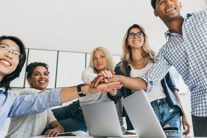 A group of five diverse people smiling and stacking hands together in a collaborative gesture. They are standing around a table with open laptops, discussing marketing strategies in a bright, modern room that opens to a sunny patio.