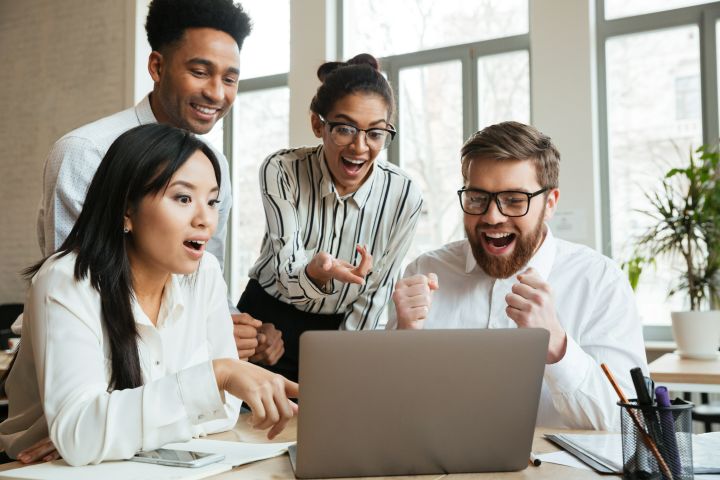 A group of four people, two men and two women, gather around a laptop with excited expressions in an office outfitted with outdoor blinds. One man raises his fists in celebration, embodying the team's success in cracking a complex marketing strategy.