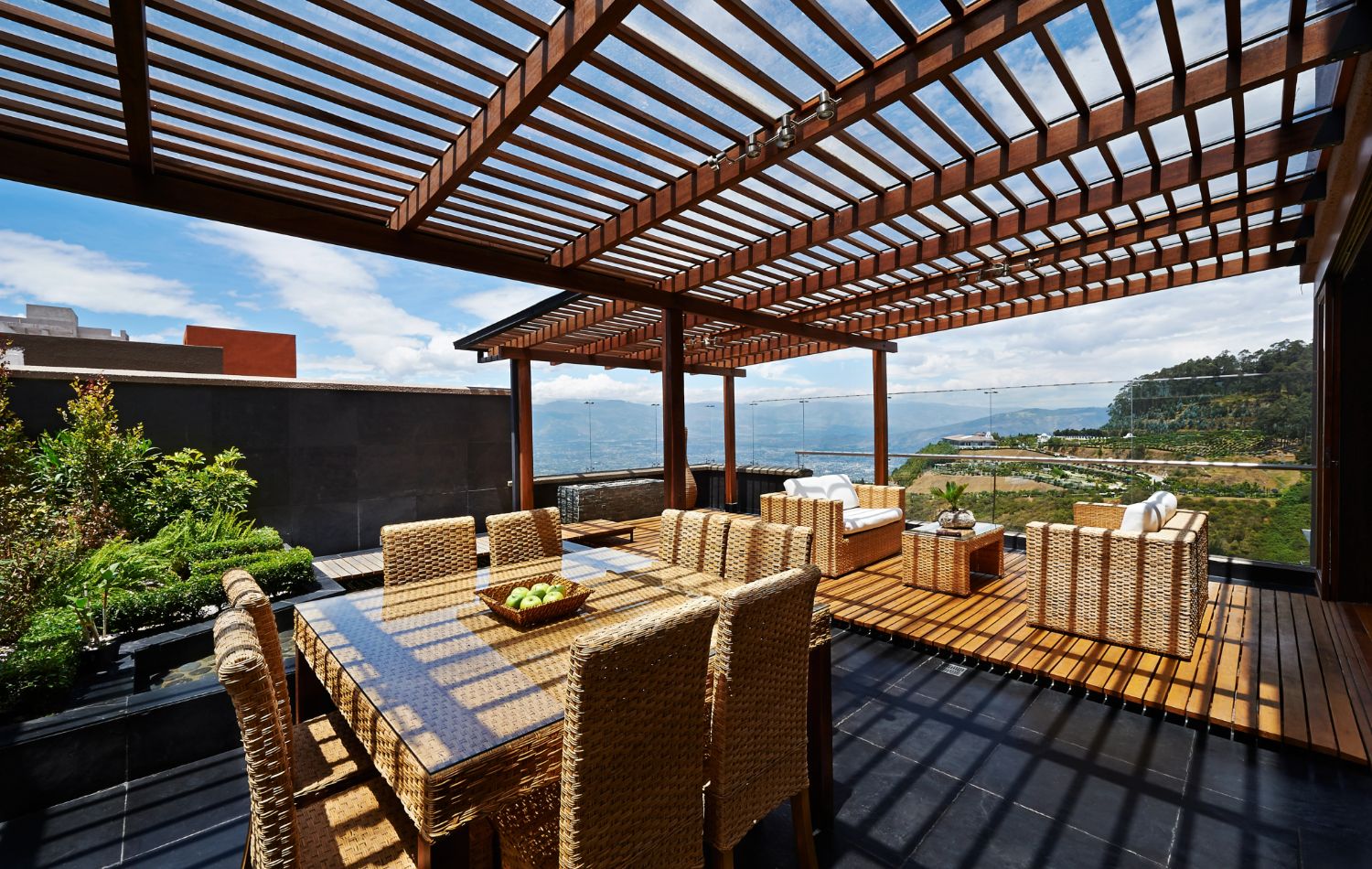 A rooftop patio with wooden pergola and outdoor dining set. Wicker chairs surround a table with decorative items. In the background, there's a lush hillside and a view of a distant water body under a clear blue sky.