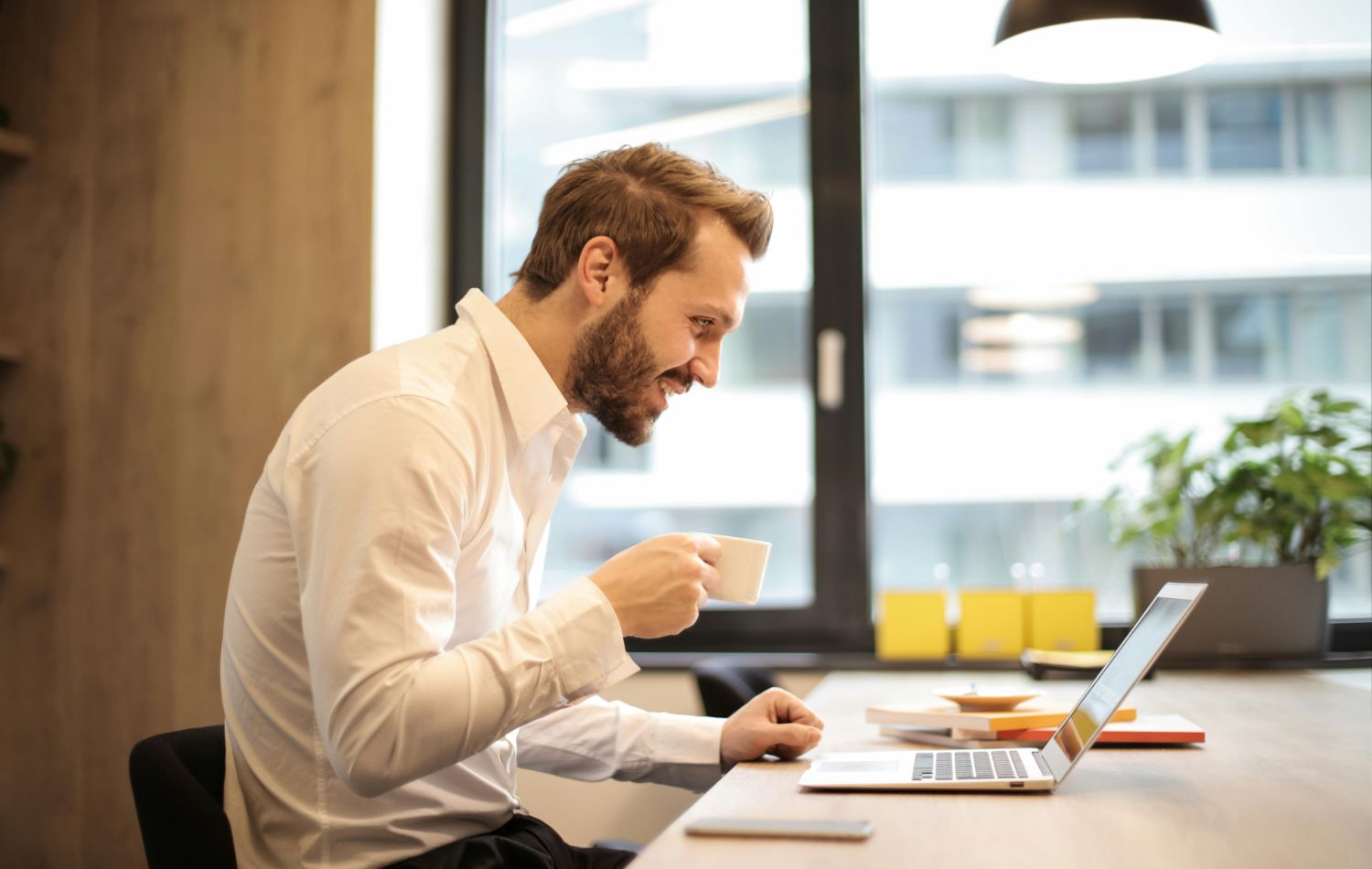 A man with a beard sits at a desk, holding a cup and looking at a laptop. He seems engaged and happy. In the background, a large window and a plant are visible. The setting appears to be an office or a workspace.