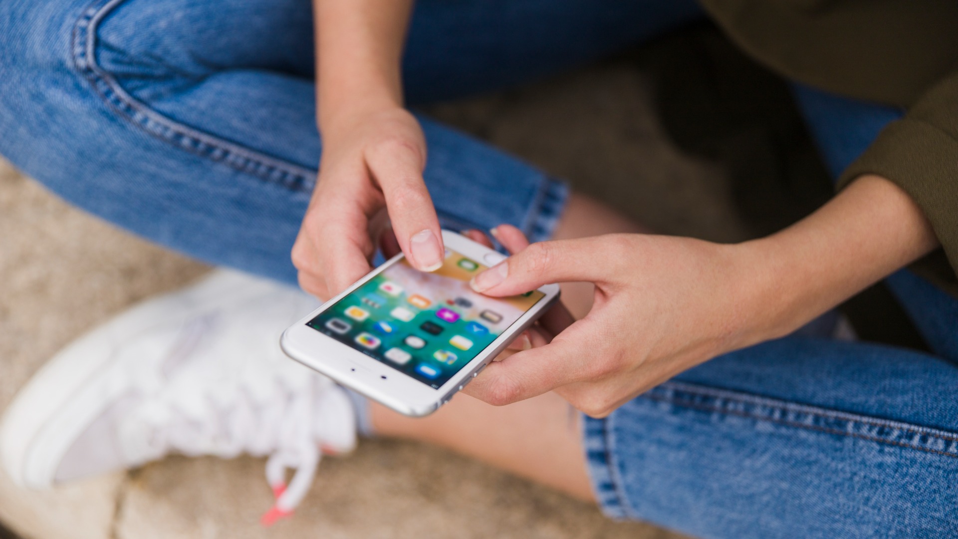 A person wearing blue jeans and white sneakers is sitting cross-legged, holding a smartphone and using their thumbs to navigate the screen.