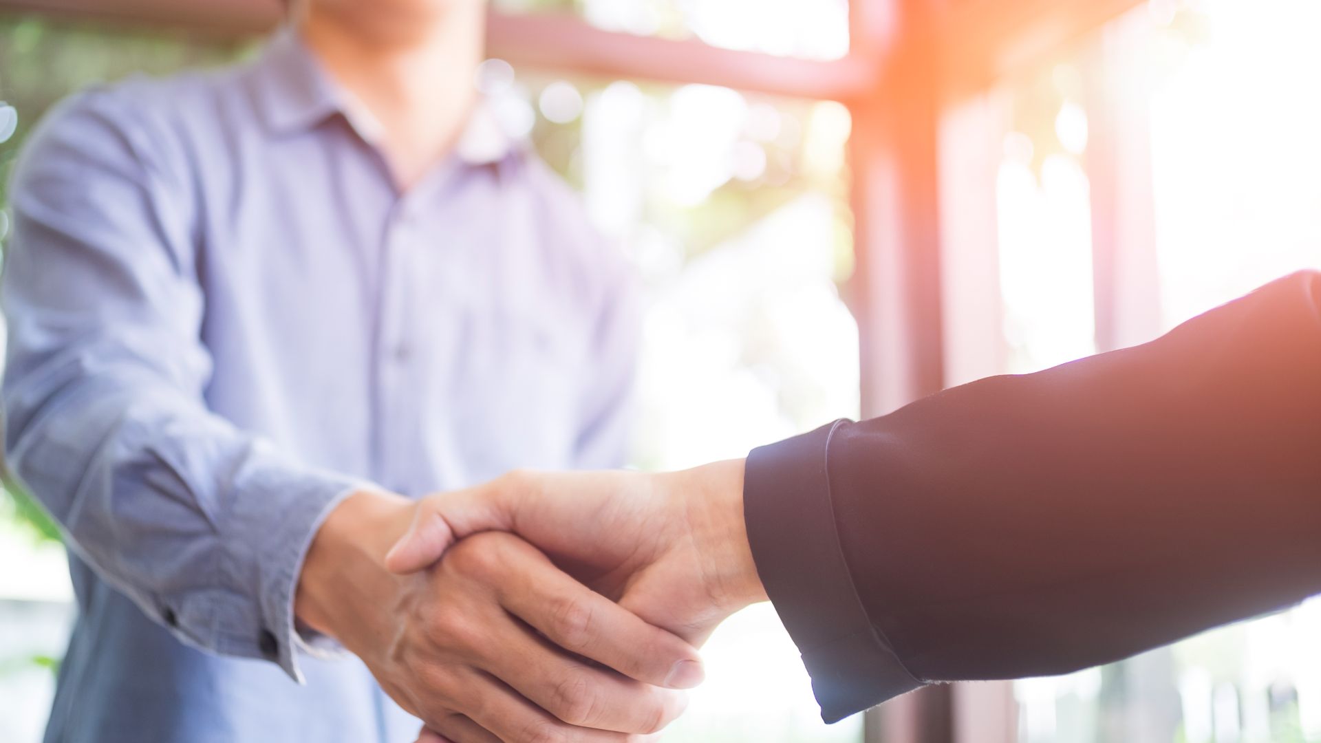 Two individuals are shaking hands in a well-lit room, reminiscent of an outdoor living space. One person is wearing a light blue shirt, while the other dons a dark suit. The scene suggests a professional setting or agreement, possibly within the realm of digital marketing strategies.