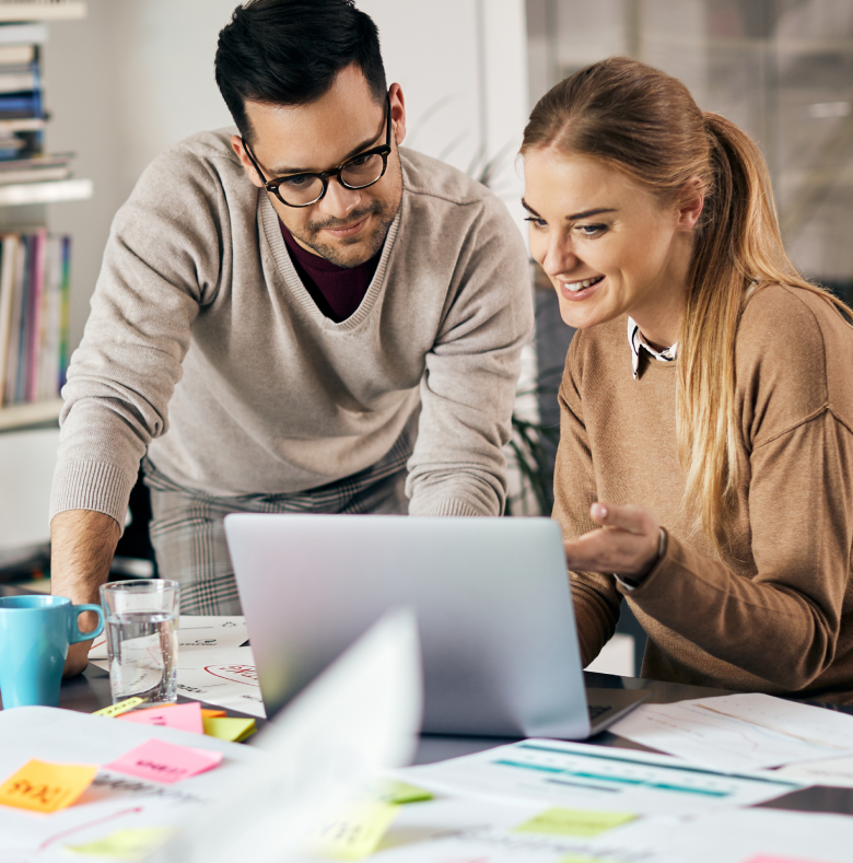 A woman and a man work together at a laptop in an office setting, immersed in discussing SEO strategies. The table is covered with papers, sticky notes, and a blue mug. They appear engaged and are both smiling. Bookshelves are visible in the background, adding to the room's studious charm.