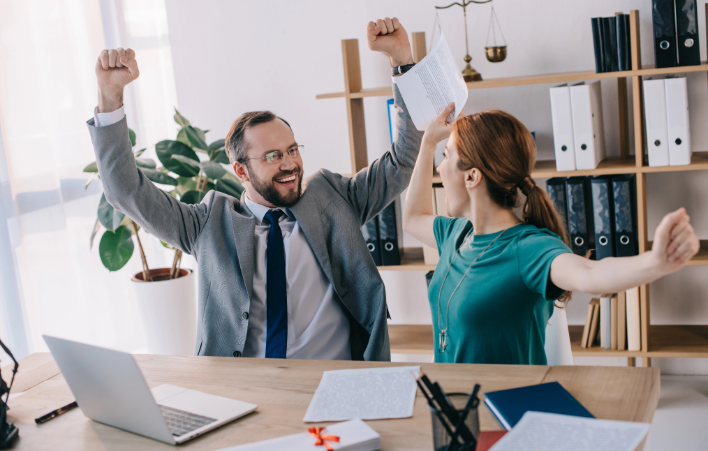 A man and woman in an office celebrate with raised arms, exuding the excitement of a successful digital marketing campaign. The man wears a gray suit and the woman a green shirt. Papers and a laptop are on the desk, with a shelf and plant in the background, enhancing their joyful atmosphere.