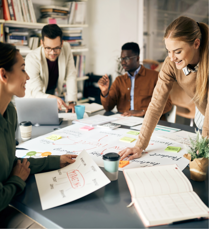 A diverse group of four people engages in a collaborative meeting, immersed in discussions on digital marketing. Gathered around a table with papers, charts, and laptops, one person stands pointing to a chart while others listen and take notes in the bright office space.