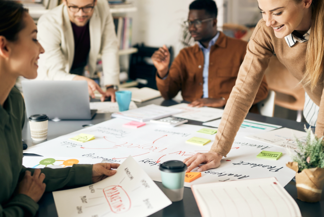 Four people collaborating around a table covered with papers, diagrams, and laptops. They are smiling and discussing digital marketing strategies, indicating an engaging and dynamic team meeting in a modern office setting.