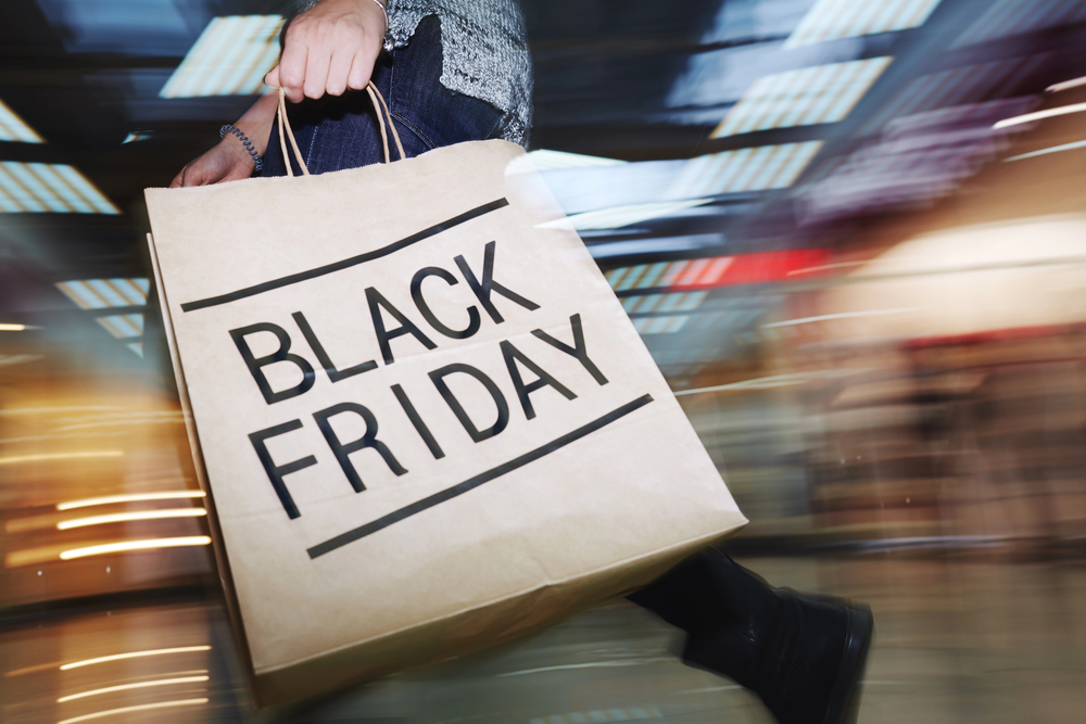 A person walking quickly through a store carrying a large shopping bag with "Black Friday" printed on it. The background is blurred, suggesting motion and urgency often associated with shopping during sales events.