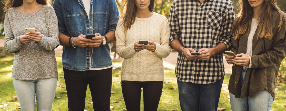 Five people standing outdoors, all looking at their smartphones. They are dressed casually in sweaters, a denim jacket, plaid shirt, and a jacket. They are standing in a line, facing forward, and the background is a park-like setting with grass and trees.