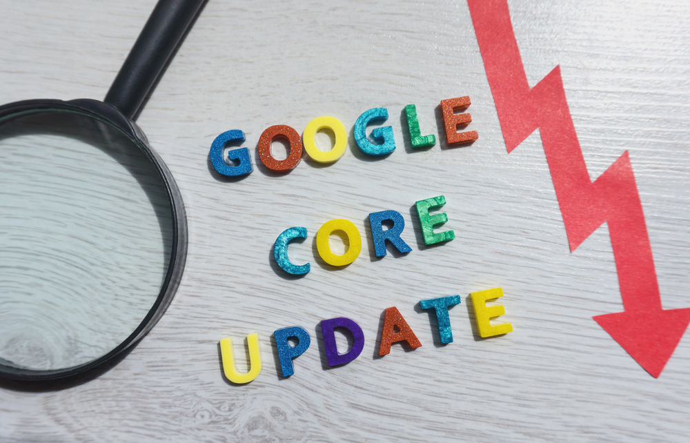 Colorful wooden letters spell out "Google Core Update" on a light wood surface. A magnifying glass is on the left, and a red zigzag arrow pointing downward is on the right.