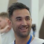 A man with short dark hair and a beard smiles, wearing a white shirt and a blue lanyard. The blurred background suggests an indoor setting with others, capturing the essence of our work environment.