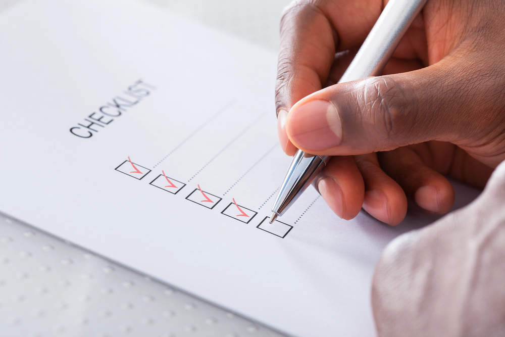 A person holds a pen and checks off a box on a checklist. The list has several completed checkboxes with red check marks. The background is a bright white surface.