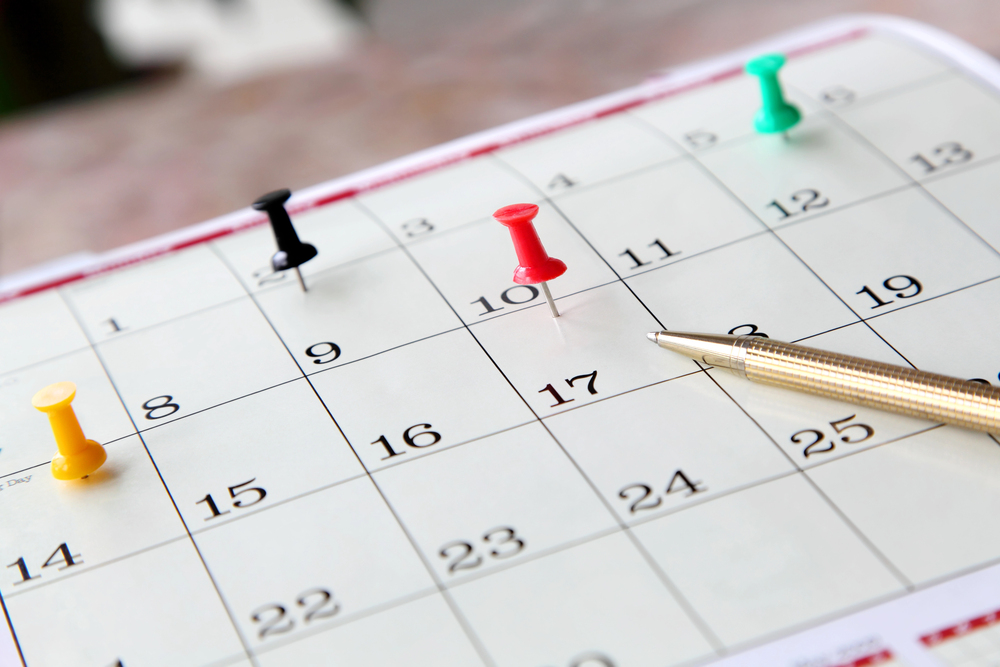 A close-up of a calendar page with push pins on the 8th, 10th, and 25th. A pen rests on the 17th, suggesting an important date. The calendar grid displays numbers, indicating days of the month.