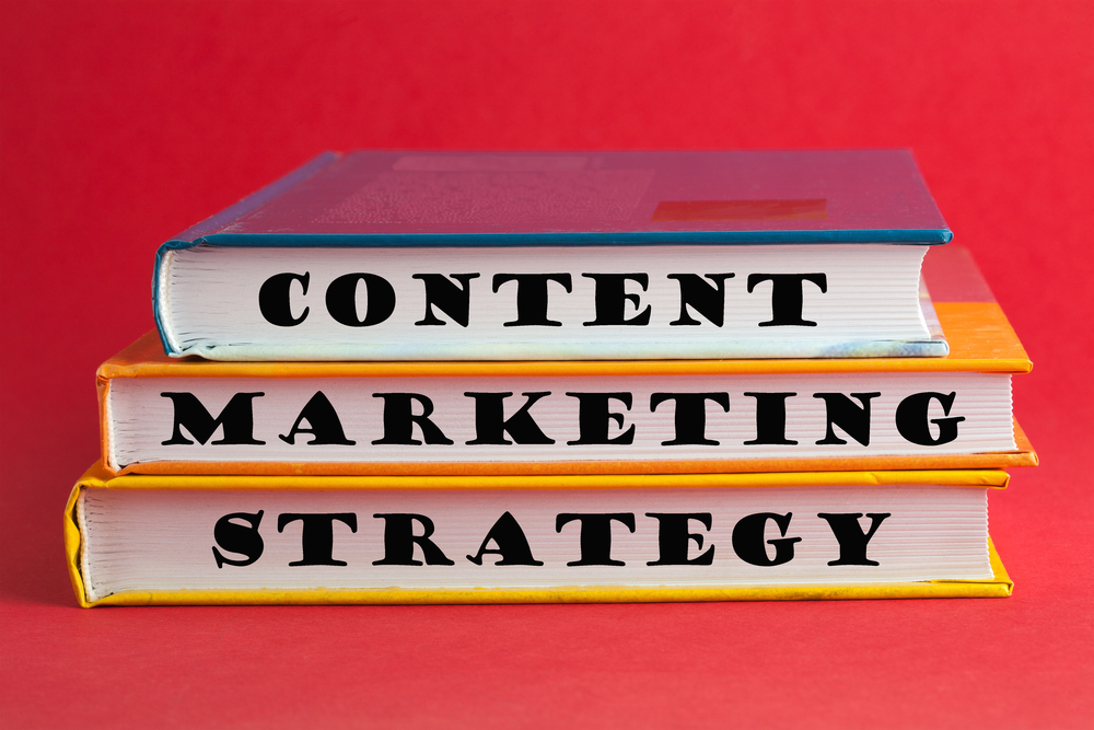 Three books stacked on a red background. The spines read "Content," "Marketing," and "Strategy," forming the phrase "Content Marketing Strategy." The books have blue, orange, and yellow covers, respectively.