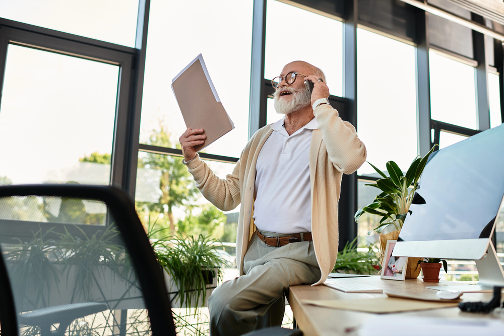 An older man with a beard, wearing glasses, stands by a desk in a modern office, holding a folder and talking on a phone. He looks engaged in conversation. The office has large windows and plants.