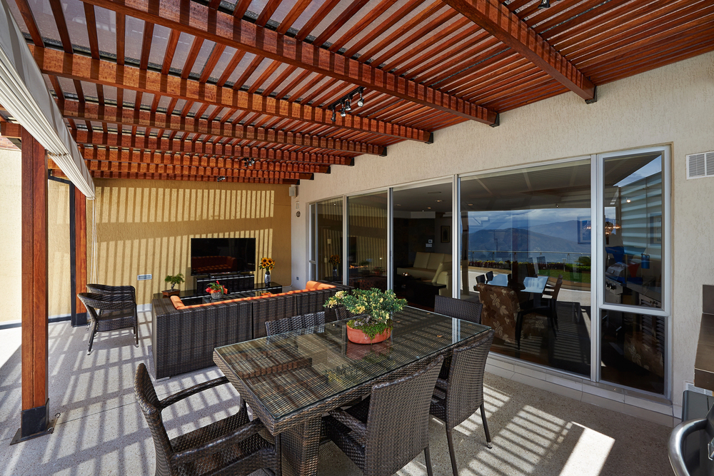 Outdoor patio with a wooden pergola, wicker table, and chairs. A potted plant sits on the table. There’s a seating area with additional wicker furniture and a wall-mounted TV. Large sliding glass doors open to reveal an interior room with mountain views.